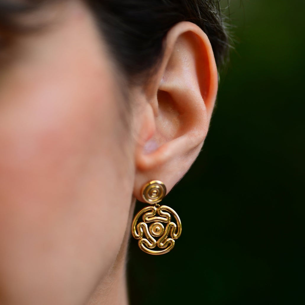 A close-up lifestyle shot of a woman wearing a gold earring. The earring features a small circular stud with a larger, detailed circular drop element in an intricate openwork pattern.