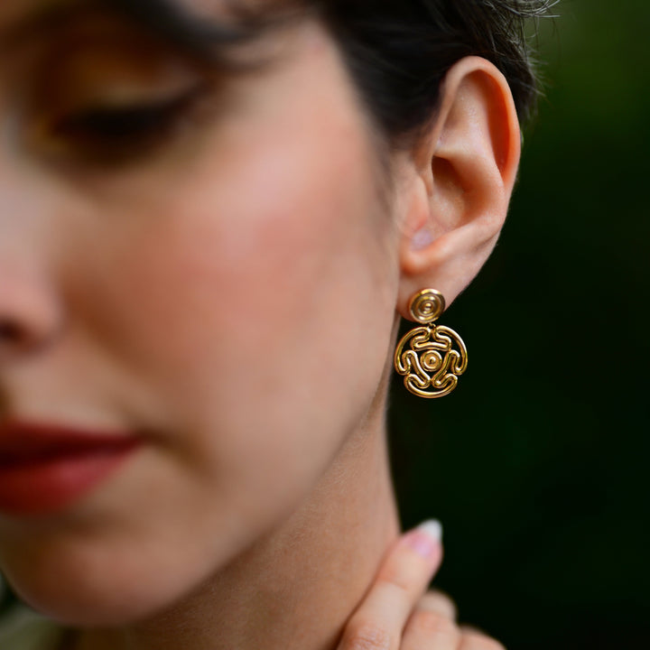 A close-up lifestyle shot of a woman wearing a gold earring. The earring features a small circular stud with a larger, detailed circular drop element in an intricate openwork pattern.