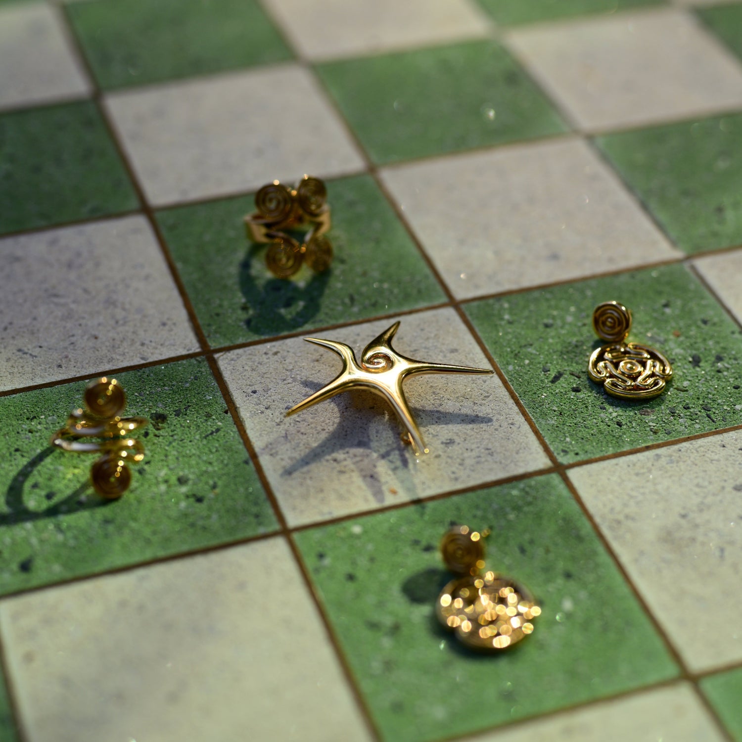 Five gold-toned earrings with organic, organic-shaped settings are arranged on a green and white checkered tile surface.