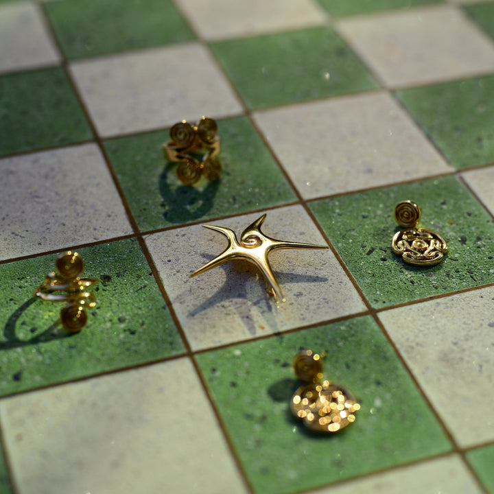 Five gold-toned earrings with organic, organic-shaped settings are arranged on a green and white checkered tile surface.