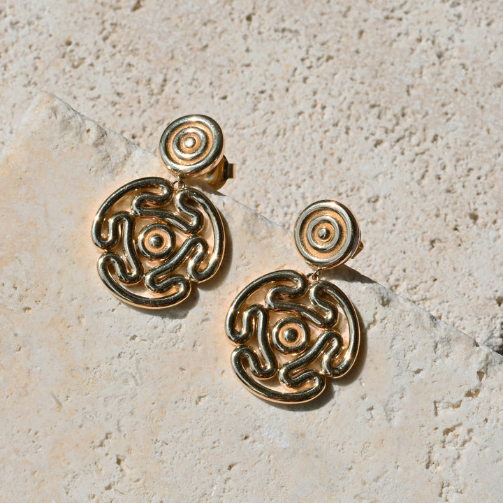 A pair of gold-toned statement drop earrings with intricate abstract spiral and loop detailing, displayed on a textured beige stone surface.