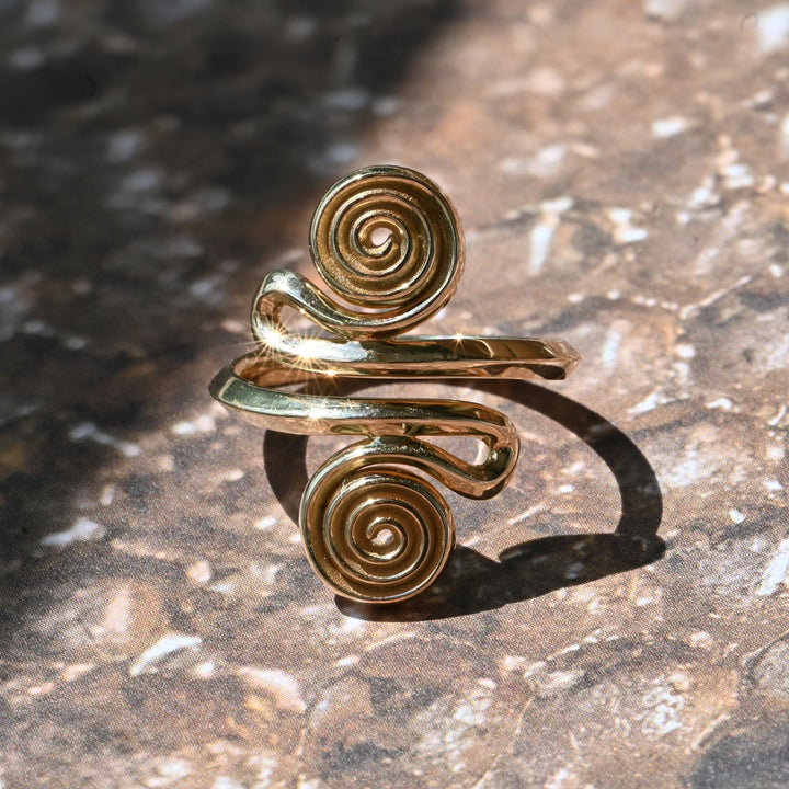 A gold spiral ring with a sculptural, organic design, resting on a textured brown stone surface under bright sunlight.
