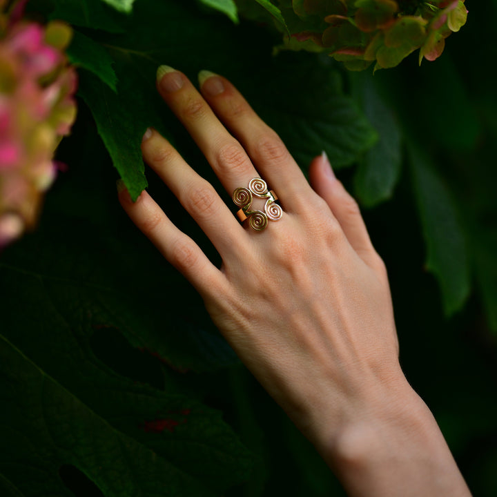 A gold ring with four spiral motifs worn on a person's hand, set against a background of dark green leaves and pink flowers.
