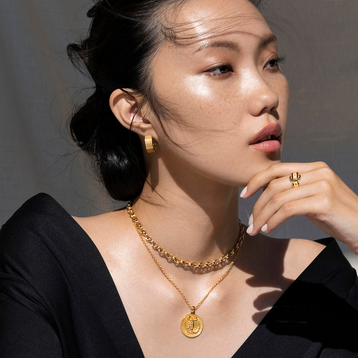 Dark Haired Woman in a Black Blouse Looking Off to the Side and Wearing Ridged Gold Hoops, a Gold Ring, and 2 Gold Necklaces