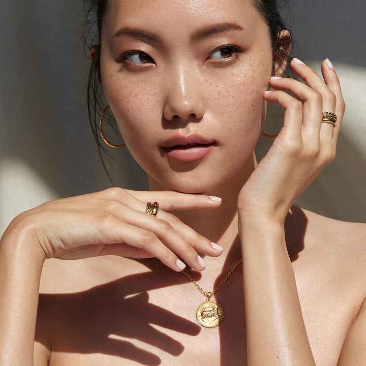 Woman Holding Her Hands by Her Face Glancing to the Side Wearing Ethical Gold Aries Pendant,  Hoop Earrings, and Band Rings