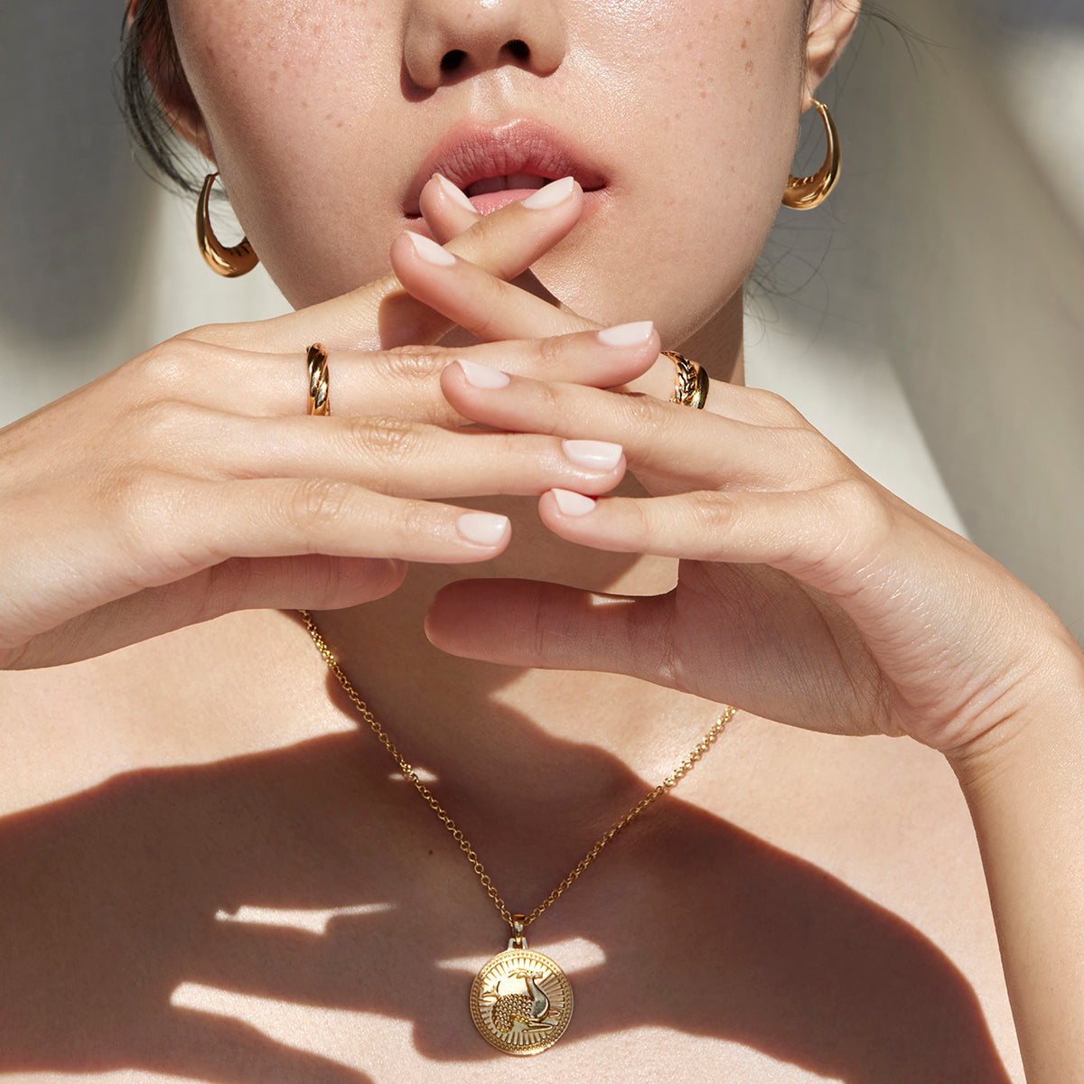 Woman with her hands tented in front of her face wearing Ethical Gold Hoop Earrings, Band Rings, and Capricorn Pendant