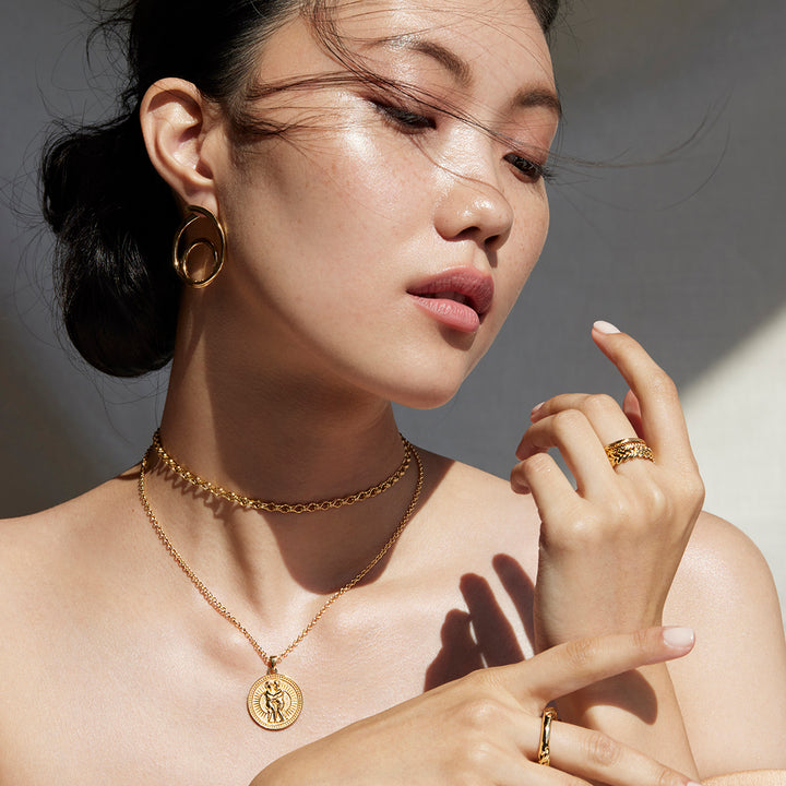 Woman Holding Her Hands by Her Face Glancing Down at Her Hands Wearing Ethical Gold Gemini Pendant, Chain Necklace, Hoop Earrings, and Band Rings