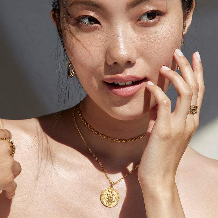 Woman Holding Her Hand by Her Face Glancing to the Side Wearing Ethical Gold Gemini Pendant, Chain Necklace, Hoop Earrings, and Band Rings