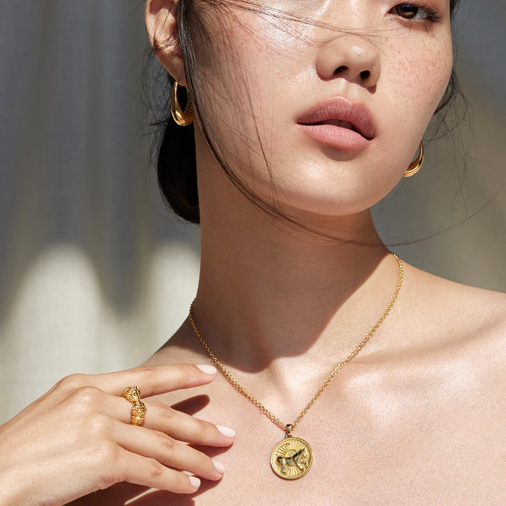 Woman Wearing Ethical Gold Leo Pendant, Lion Ring, and Hoop Earrings Holding Her Hand by Her Collar Bone