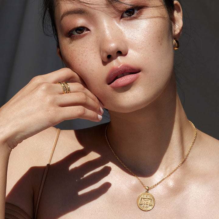 Woman Facing the Camera Holding a Hand Against Her Chin Wearing Libra Ethical Gold Pendant and Ethical Gold Band Rings