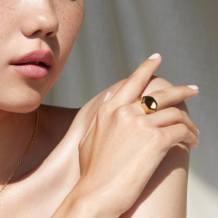 Woman with Her Hand Over Her Shoulder Wearing a Sustainable Gold Ring Featuring a Flat Hexagonal Design at the Top of the Ring on her Middle Finger